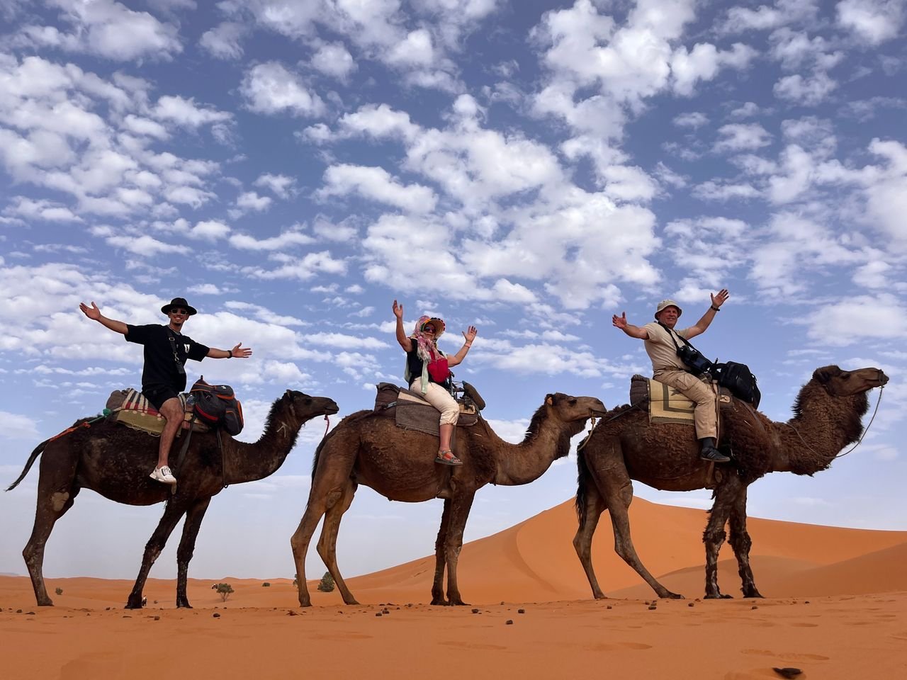 Three tourists riding camels in the Sahara Desert under a bright blue sky.